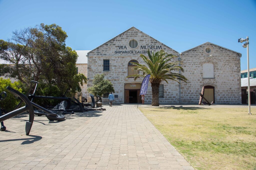 Front entrance of WA Shipwrecks Museum in Fremantle, Western Australia, with palm trees, historic limestone building, and large ship anchors on display.