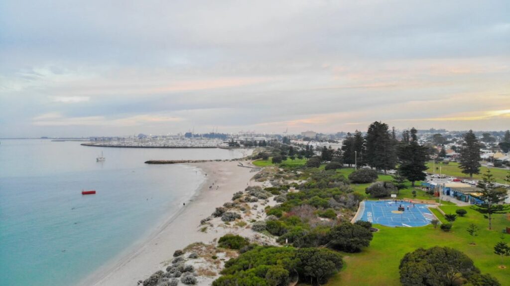 Aerial view of South Beach Reserve in Fremantle with white sandy shoreline, turquoise water, basketball courts, and lush green parklands at sunset.