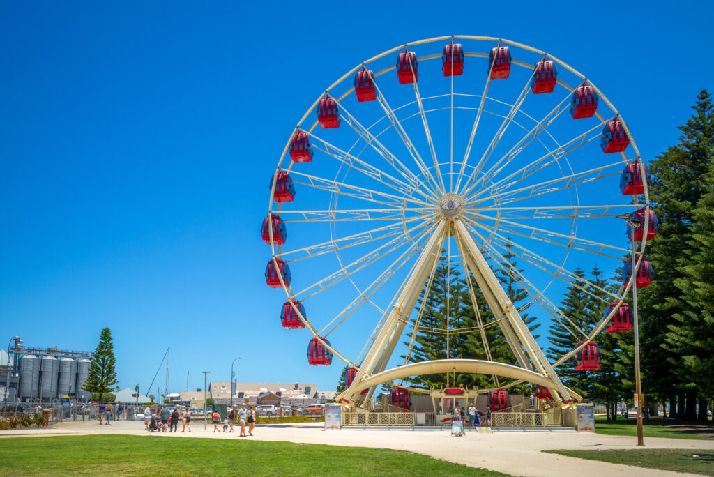 Fremantle Tourist Wheel at Esplanade Park on a sunny day, with people walking nearby and tall pine trees in the background.
